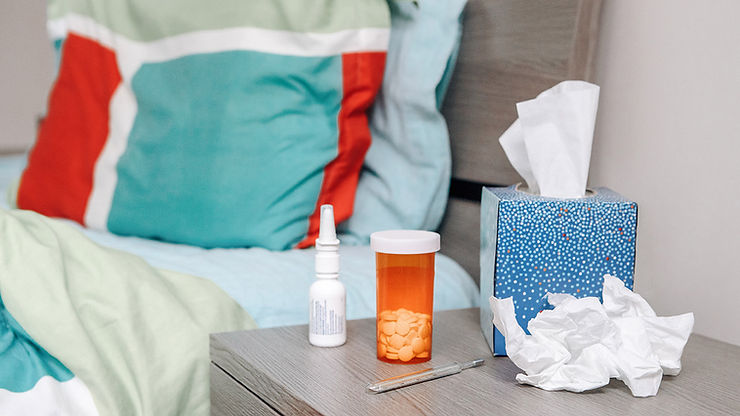 A view of a bedside table with a tissue box, pill bottle, nasal spray, and thermometer. A bed is visible in the background.