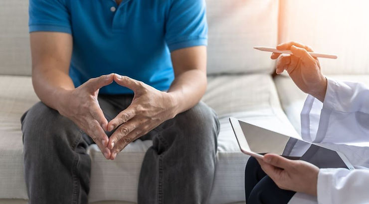 A man in a blue shirt and gray pants sits on a white couch beside a doctor in a white coat, holding a pencil and an iPad.