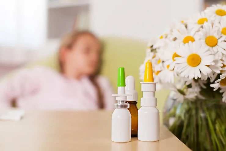 A tan table has three bottles, brown and white with yellow and green tips, on it beside a vase of daisy flowers, with a woman lying down in the background.