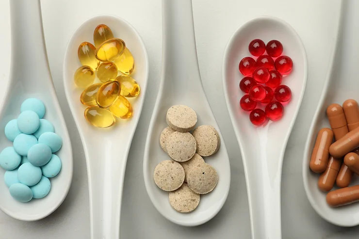 Five white spoons are placed side by side in front of a white background, each holding blue, yellow, brown and red pills.
