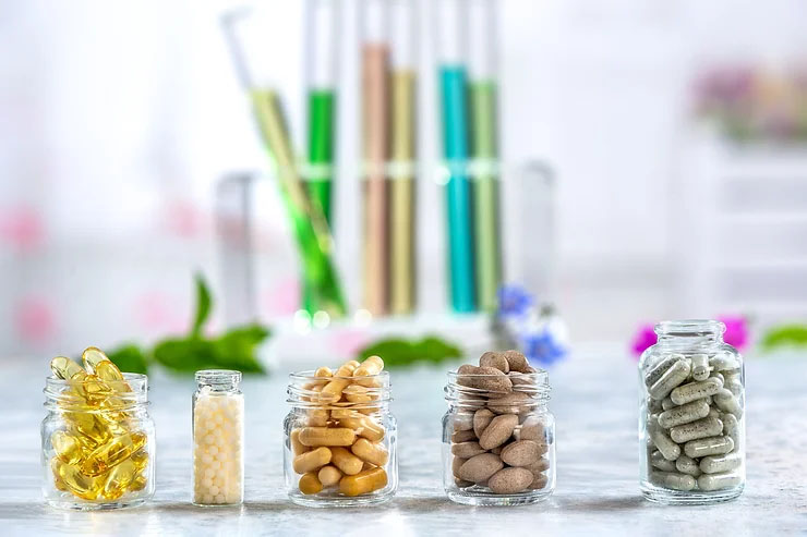 Five glass bottles sit side by side on a white countertop, filled with yellow, brown, and gray pills with vials of yellow, green, brown, and blue fluids.