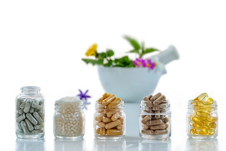 Five glass jars filled with green, white, brown, and yellow pills in front of a white bowl filled with pink and yellow flowers.