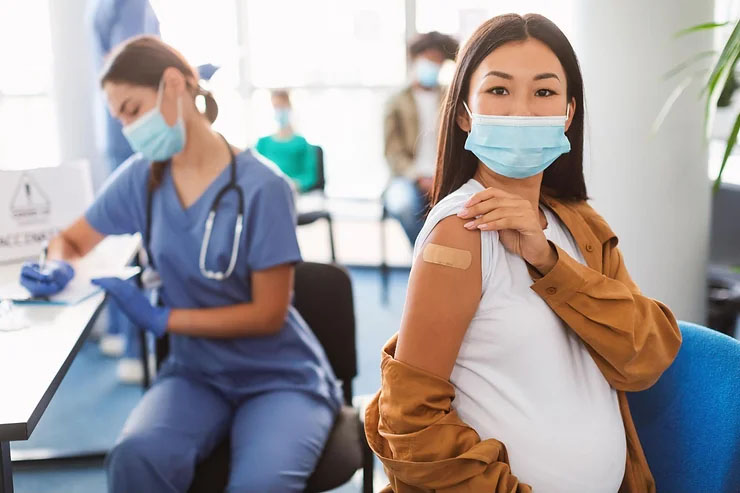 A smiling pregnant woman with a face mask and her brown jacket lowered shows her arm with a vaccine patch while a doctor records the vaccination.