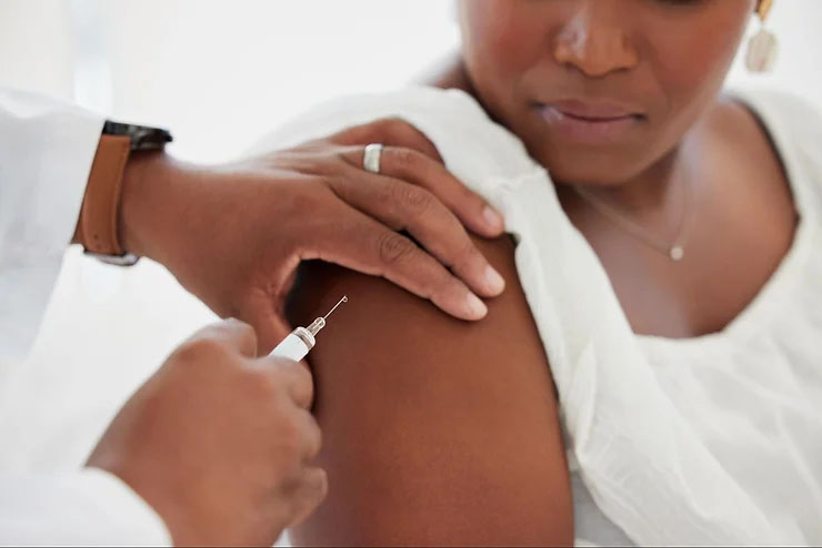 An African American patient in a white tank top receiving a vaccine injection with a close-up of the doctor's hand holding a syringe.