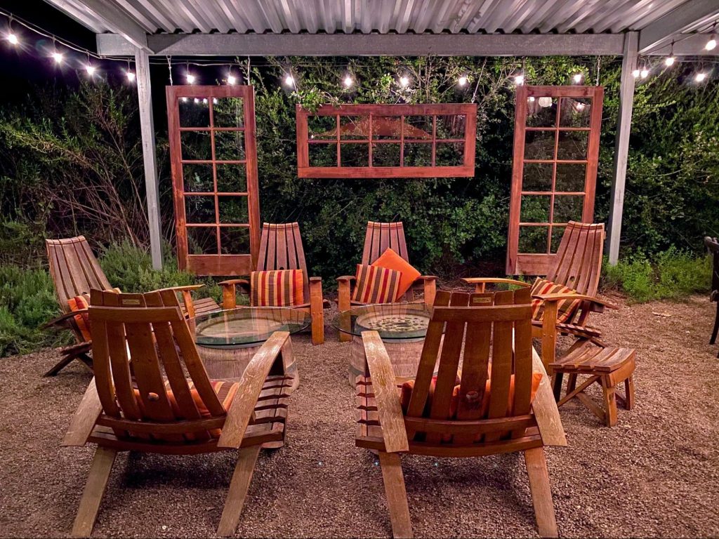 A covered outdoor living space at night with hanging wooden door frames adorned with string lights, two round glass tables in the center, Mexican-inspired throw pillows, and lush greenery in the background.  