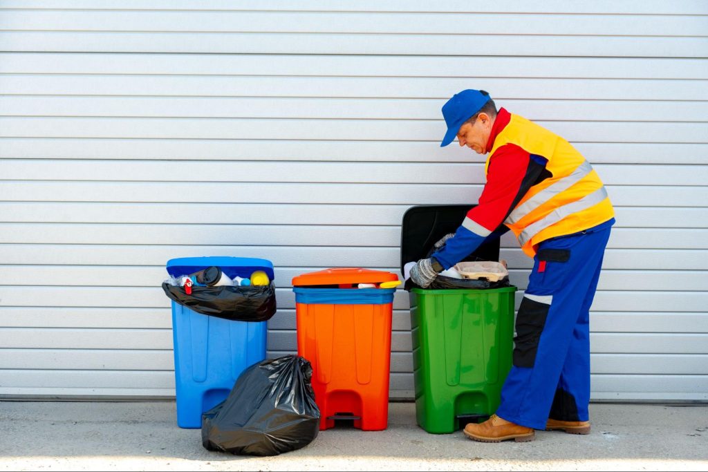 A janitor in a vibrant red, blue, and yellow uniform removes garbage from a multi-colored trash container outdoors, demonstrating professional trash pick-up services in action.
