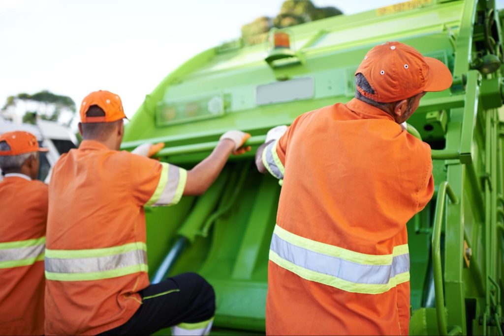Three workers in orange uniforms next to a trash truck.